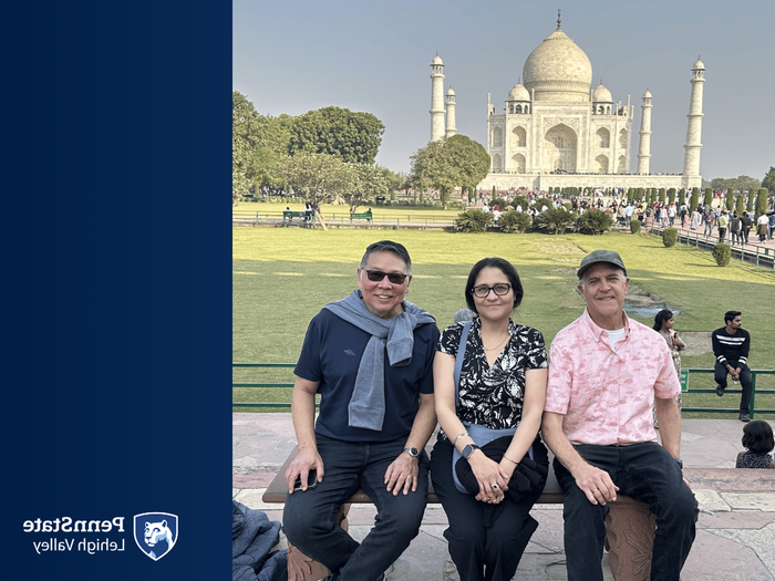 two men and a woman sit on a bench and smile in front of the Taj Mahal in India